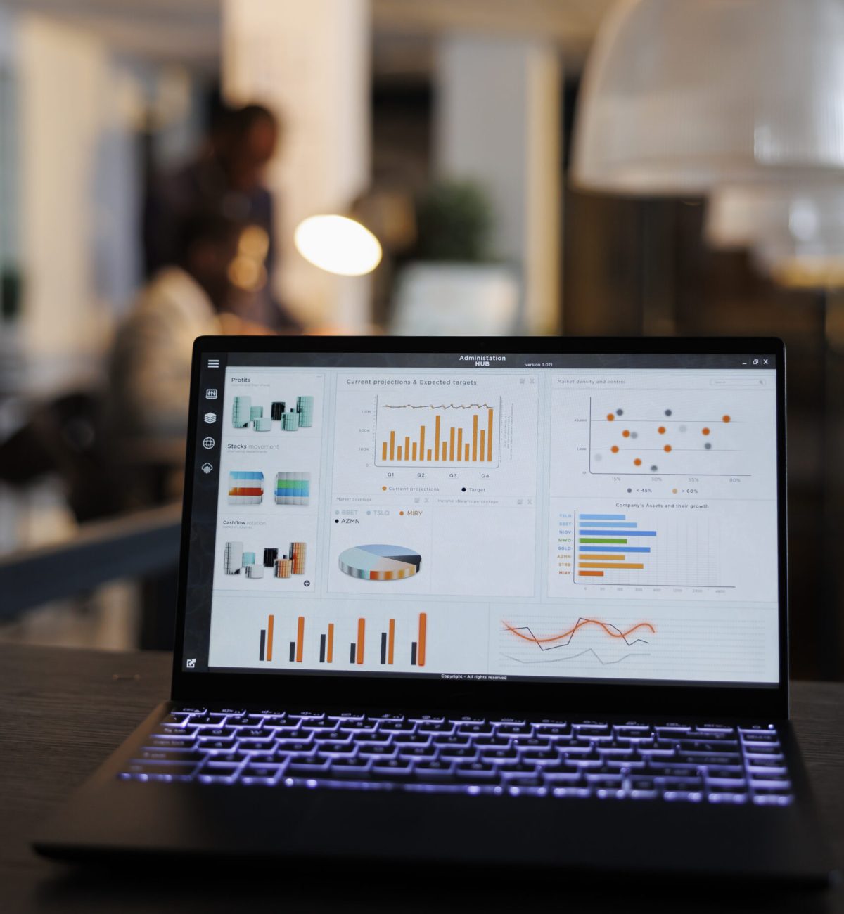 Laptop computer with marketing statistics graphs on screen, standing on table in startup office, coworkers discussing company strategy in background. Employees working overtime in startup office
